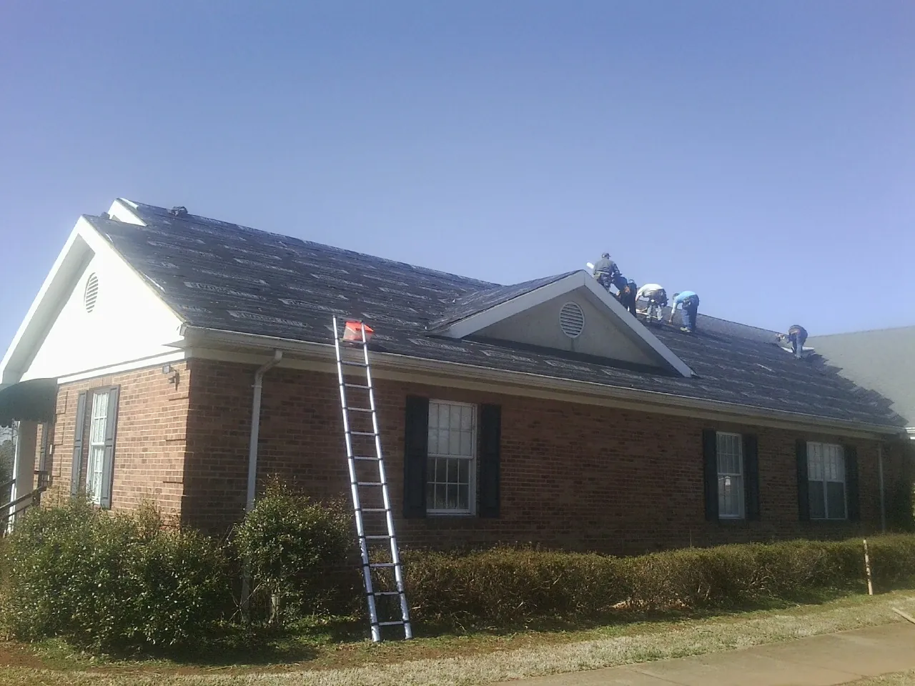 Crew tearing shingles off a brick ranch roof with a ladder set — Emergency Roof Repair work by our crew in Mandaree