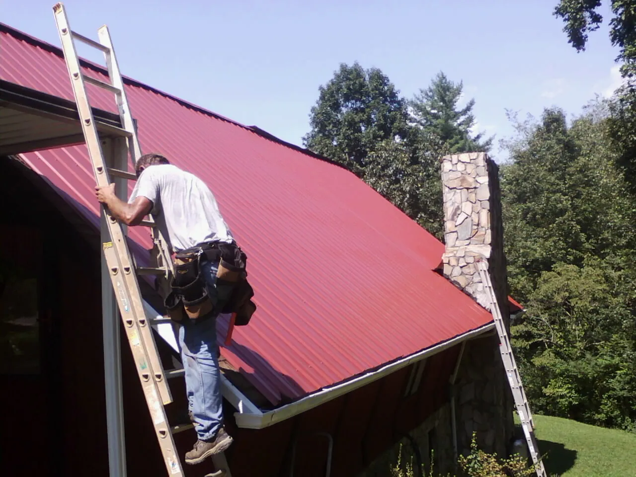 Roofer on a ladder beside a red metal roof and stone chimney — Soffit & Fascia Repair by Roofing Brothers in Mandaree